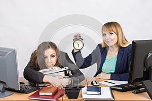 Two girls in the office at the end of the day, one with a smile, holding a clock, another weary lies on folders