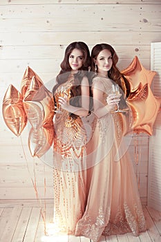 Two Girls on party. Beautiful young women in elegant golden dress with champagne glasses over golden star balloons background. H