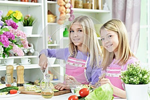 Two girls in pink aprons preparing salad on kitchen table