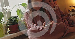 Two girls in pink dresses are sitting in the kitchen and playing games on the phone.