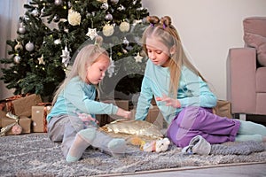 Two girls sisters draw fingers on pillow with sequins sit near Christmas tree.