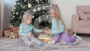 Two girls sisters draw fingers on pillow with sequins sit near Christmas tree.