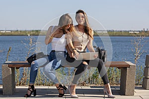 Two girls sit on a bench in the summer park. They look at the laptop screen