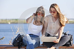 Two girls sit on a bench in the summer park. They look at the laptop screen