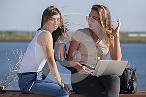 Two girls sit on a bench in the summer park. They look at the laptop screen