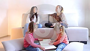 Two girls sit on floor and flip book, young moms sit on sofa next to and discuss problem.