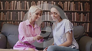 Two girls sit in the library and read a book in electronic form