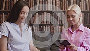 Two girls sit in the library and read a book in electronic form