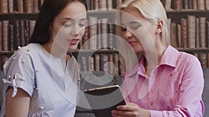 Two girls sit in the library and read a book in electronic form