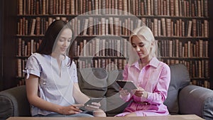 Two girls sit in the library and read a book in electronic form