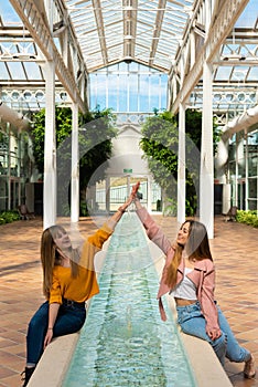 Two girls sitting on the edge of a fountain with water inside a bright greenhouse