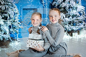 two girls are sitting near the Christmas tree. red-haired sisters unfold gifts near the fireplace. the interior is blue