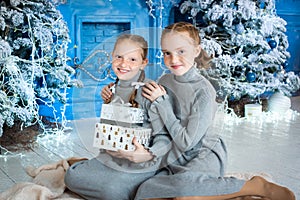 two girls are sitting near the Christmas tree. red-haired sisters unfold gifts near the fireplace. the interior is blue