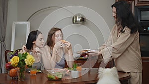 Two girls are sitting at a table in the living room and eating fresh vegetable salad and fruit. A third girl approaches