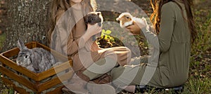 Two girls are sitting under a tree on the grass and petting little rabbits, holding a large gray rabbit in a box nearby. Pastel