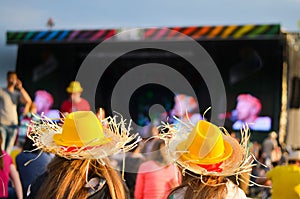 Two girls on summer festival sitting in front the stage and listening music with yellow hats on the head
