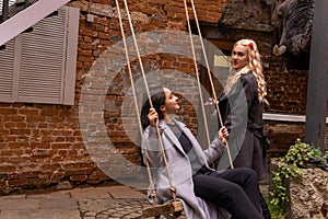 Two girls are talking in the courtyard of an old building, one of them is sitting on a swing