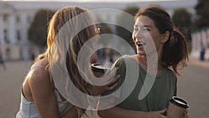 Two girls teenage friends meet each other in street european town laughing at something outside and drink coffee. slow