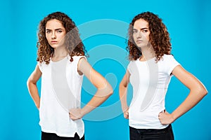 Two girls twins posing with arms akimbo over blue background.