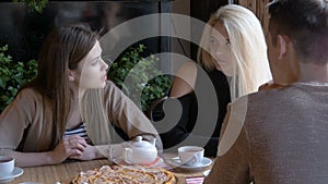 Two girls and a young man talking in a cafe sitting at a table. They drink tea and eat pizza. They tell the news, they