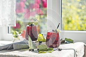 Two glass of cold ice black currant juice with ripe berries and green leaves on table in sunny room near window with garden