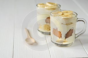 Two glass cups with banana pudding and wooden spoon on a white table. Milk and banana dessert.