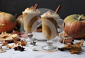 Two glass cups pumpkin latte with spices on grey background with pumpkins and autumn leaves, close-up.