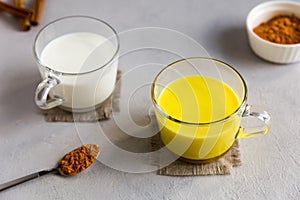 Two glass cups stand on gray background.One ordinary milk,other golden milk drink with turmeric root powder.