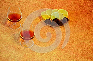Two glass glasses with cognac and a plate with chocolates and lemon slices on a brown background