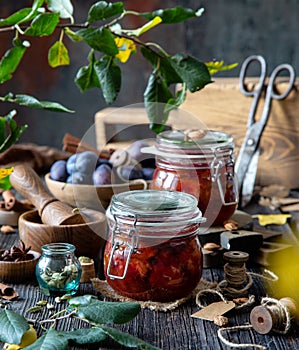 Two glass jars with homemade canned plums jam, marmalade, jelly on rustic wooden table with cardamon, cinnamon,  anise, plums