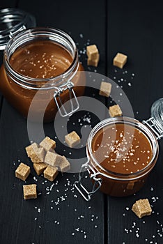 Two glass jars with homemade salted caramel and pieces of brown sugar with salt crystals