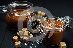 Two glass jars with homemade salted caramel and pieces of brown sugar with salt crystals