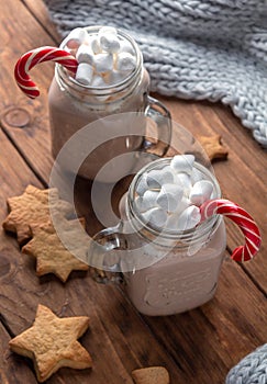 Two glass mugs with cocoa, marshmallows, sugar cane and ginger cookies on a wooden background.