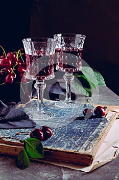 Two glass with a red alcoholic drink, old books and ripe cherry berries on a black background. Still life with red wine, ripe