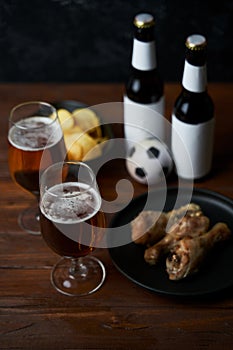 Two glasses with beer, bottles, snacks and soccer ball on wooden table.