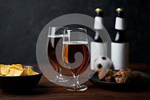 Two glasses with beer, bottles, snacks and soccer ball on wooden table.