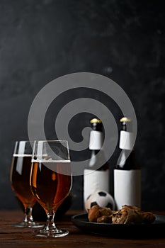 Two glasses with beer, bottles, snacks and soccer ball on wooden table.