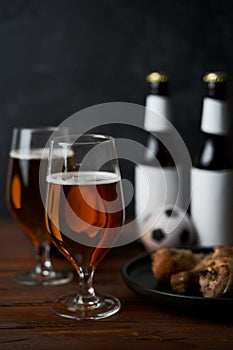 Two glasses with beer, bottles, snacks and soccer ball on wooden table.