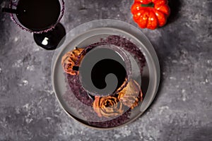 Two glasses with black cocktail, dried roses, pumpkin for Halloween party on the dark background