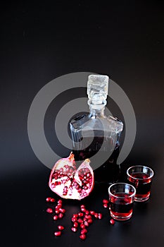 Two glasses with bright red liqueur and a glass bottle on a black background, next to a ripe pomegranate and fruit seeds