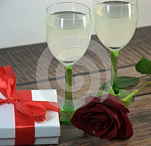 Two glasses of champagne, red rose and gift box on a wooden table