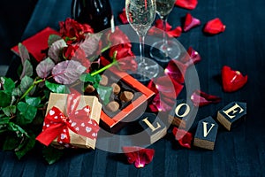 Two glasses of champagne, red roses, petals, gift box with red ribbon, chocolates and wooden love words on a black background