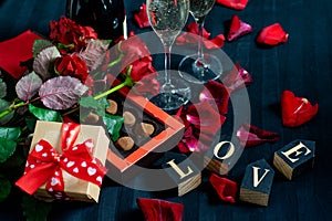 Two glasses of champagne, red roses, petals, gift box with red ribbon, chocolates and wooden love words on a black background