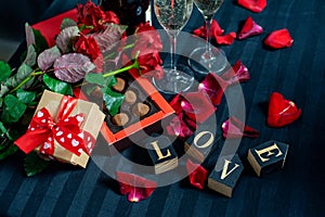 Two glasses of champagne, red roses, petals, gift box with red ribbon, chocolates and wooden love words on a black background