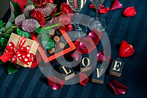 Two glasses of champagne, red roses, petals, gift box with red ribbon, chocolates and wooden love words on a black background