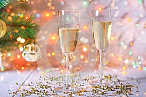 Two glasses of champagne stand on a white wooden table on the background of a New Year tree and garlands. Christmas bokeh.