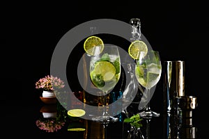 Two glasses with cold drink decorated with lemon mint and ice, with bottles of booze and cocktail shaker