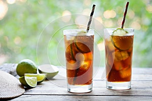 Two glasses Cuba Libre cocktail with a cola, ice, lime and rum on wooden table with a view to the terrace. Close up