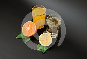 Two glasses with different citrus juices on a black background, next to half a ripe lemon and orange with leaves