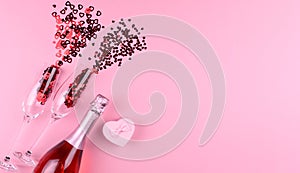 Two glasses filled with heart sequins with a bottle of champagne and a Valentine`s Day gift on a pink background